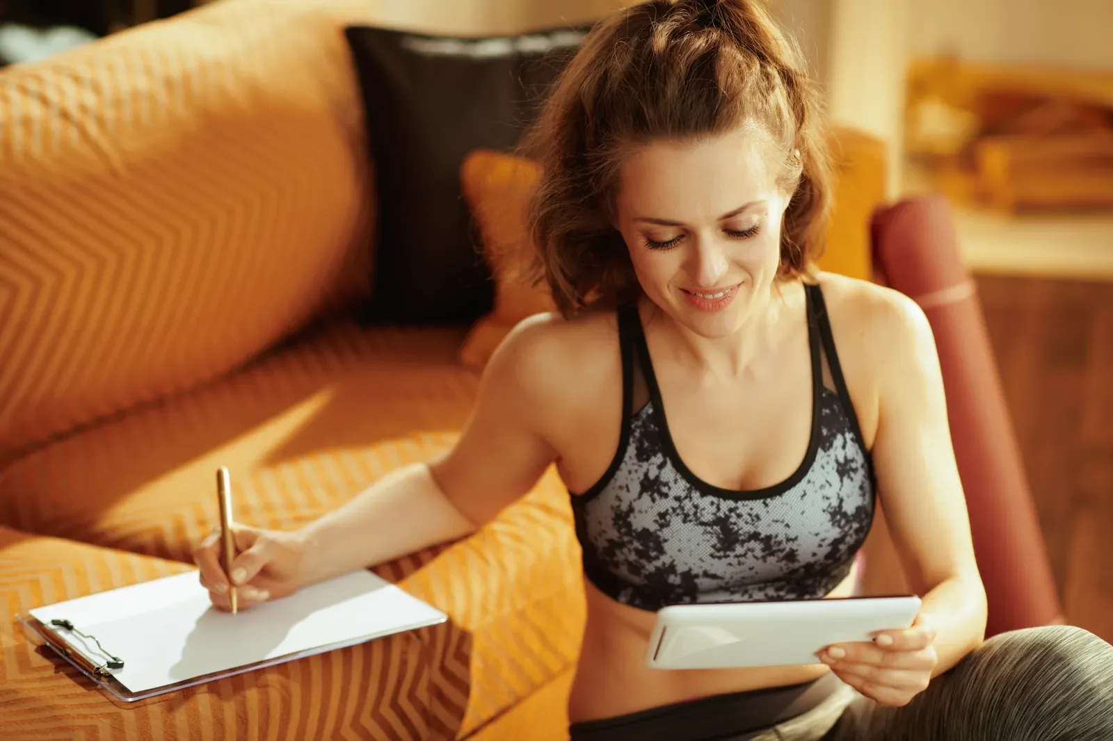 Fitness coach at home writing a session plan on a clipboard while holding a tablet